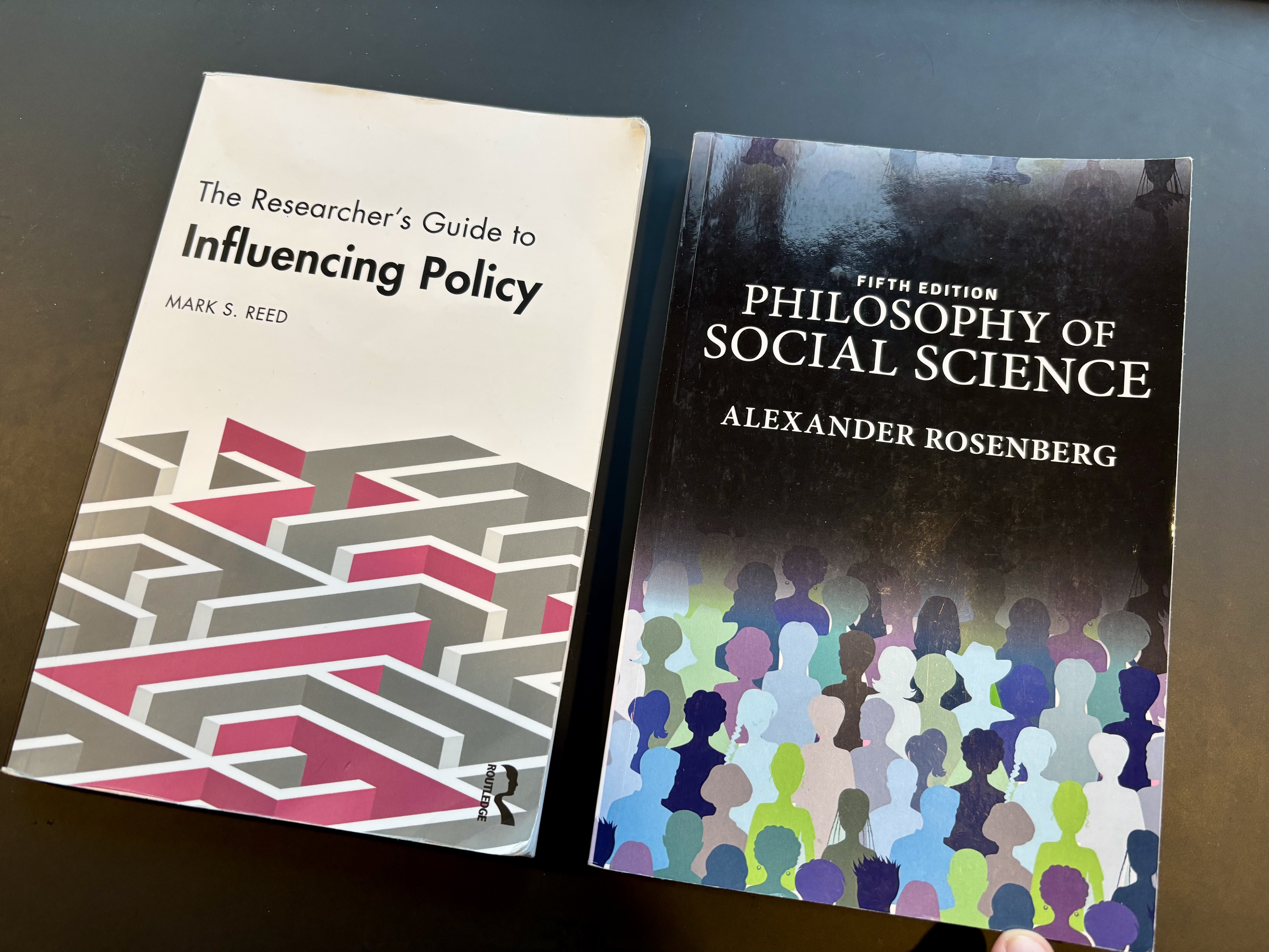 A picture of two books on a dark countertop. The book on the left is titled The Researcher's Guide to Influencing Policy. The book on the right is titled Philosophy of Social Science.